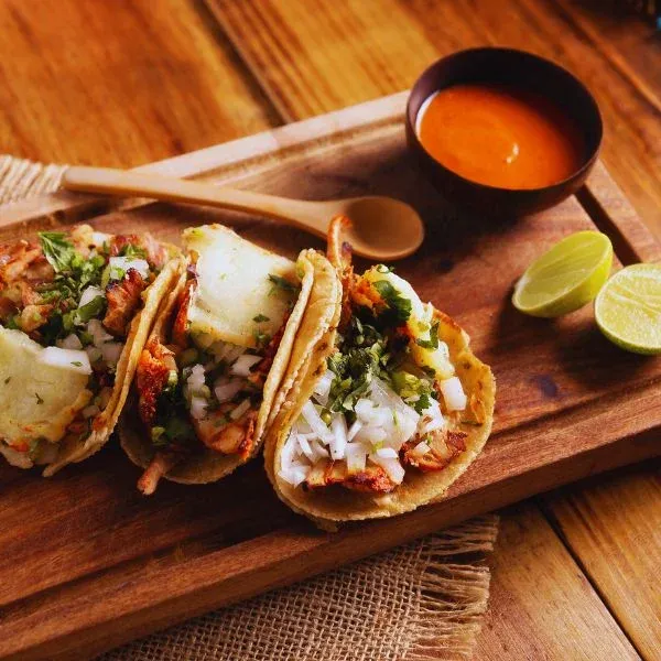 Trois tacos al pastor garnis de coriandre et d'oignon, sauce et citron vert sur une planche
